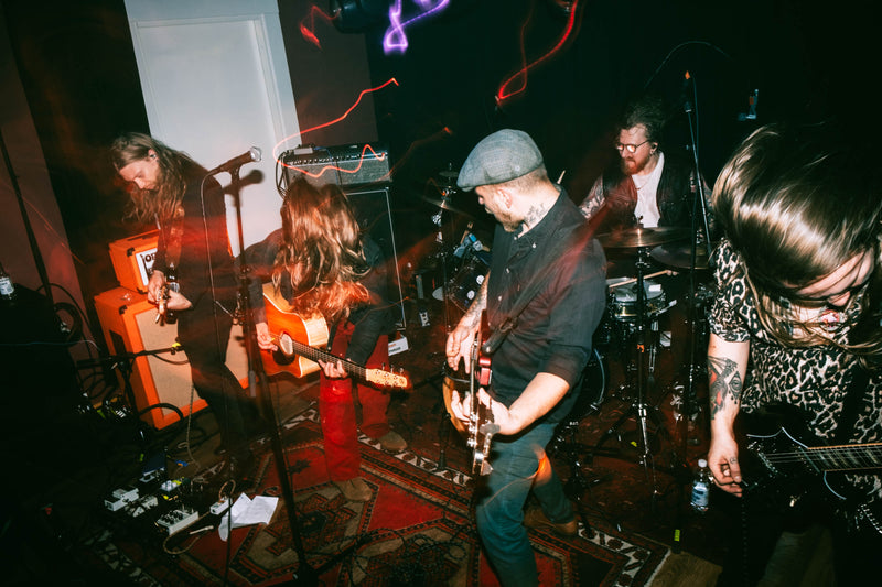 A live band performing on stage in a dark venue, with multiple musicians playing guitars, a bass, drums, and amplifiers under colored stage lighting.