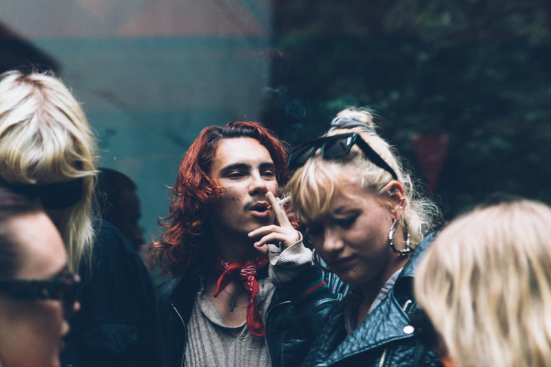 A candid street-style photograph featuring two young adults in a crowd. The person on the left has wavy red hair, a red bandana, and a gray knit sweater, holding a cigarette.