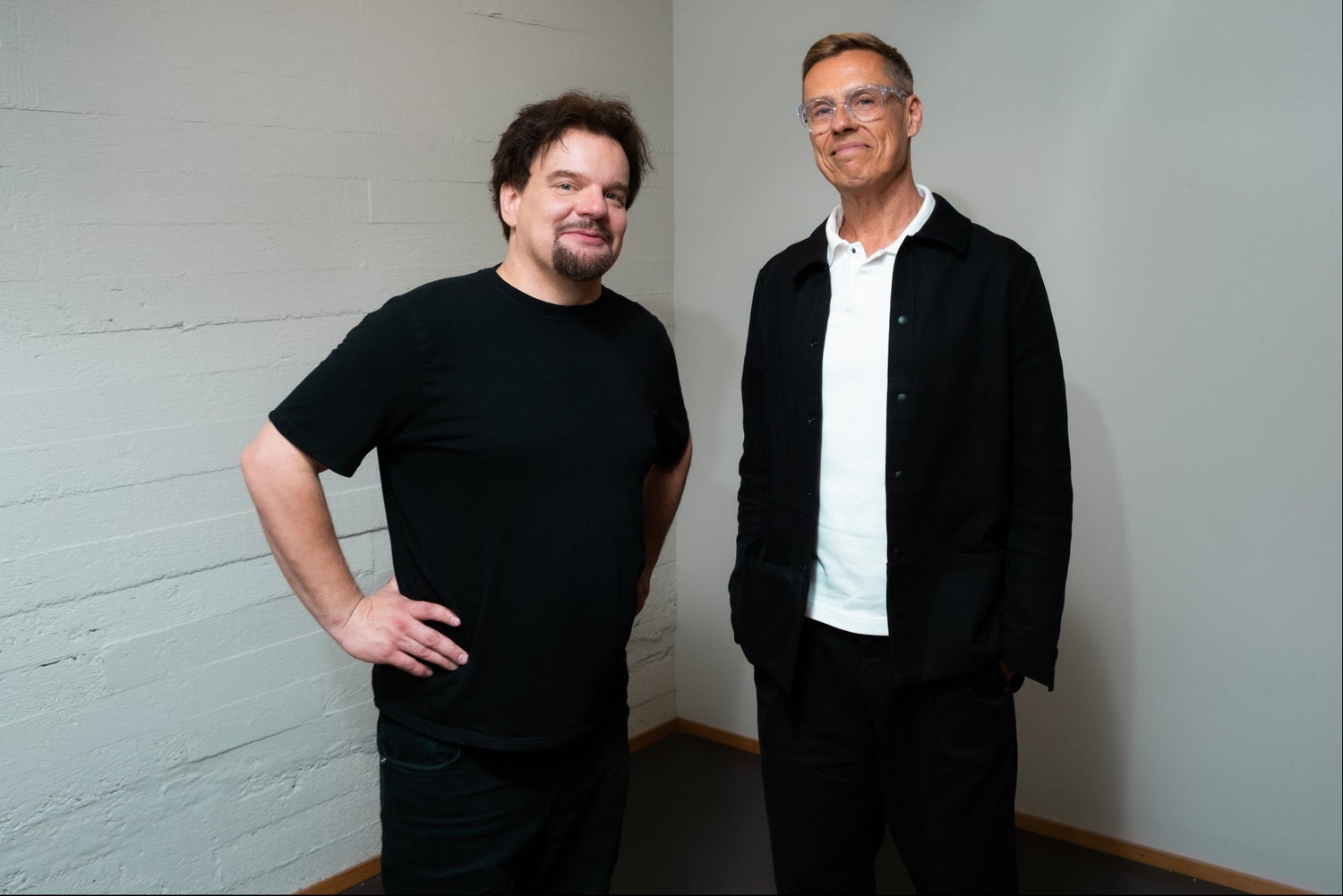 Alexander Stubb and Ismo Leikola Helsinki, Finland. Two men standing in a studio with white brick walls. The man on the left wears a black t-shirt and black pants, while the man on the right wears glasses, a black jacket over a white collared shirt, and black trousers.