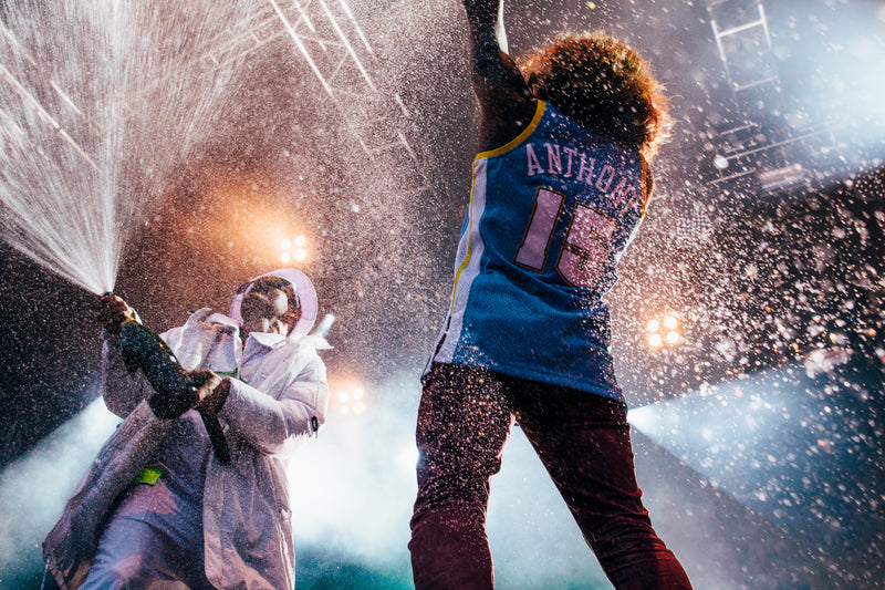 Pete Parkkonen JVG. A person in a blue and yellow basketball jersey with 'ANTHONY 18' on the back, standing under stage lights while water sprays heavily across the scene. Another person in a white hooded jacket is visible spraying water on the left.