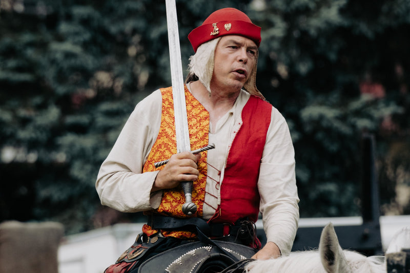 A man in medieval-style costume riding a white horse outdoors, wearing a red hat, red vest, patterned orange vest, and black pants, while holding a sword. turku finland