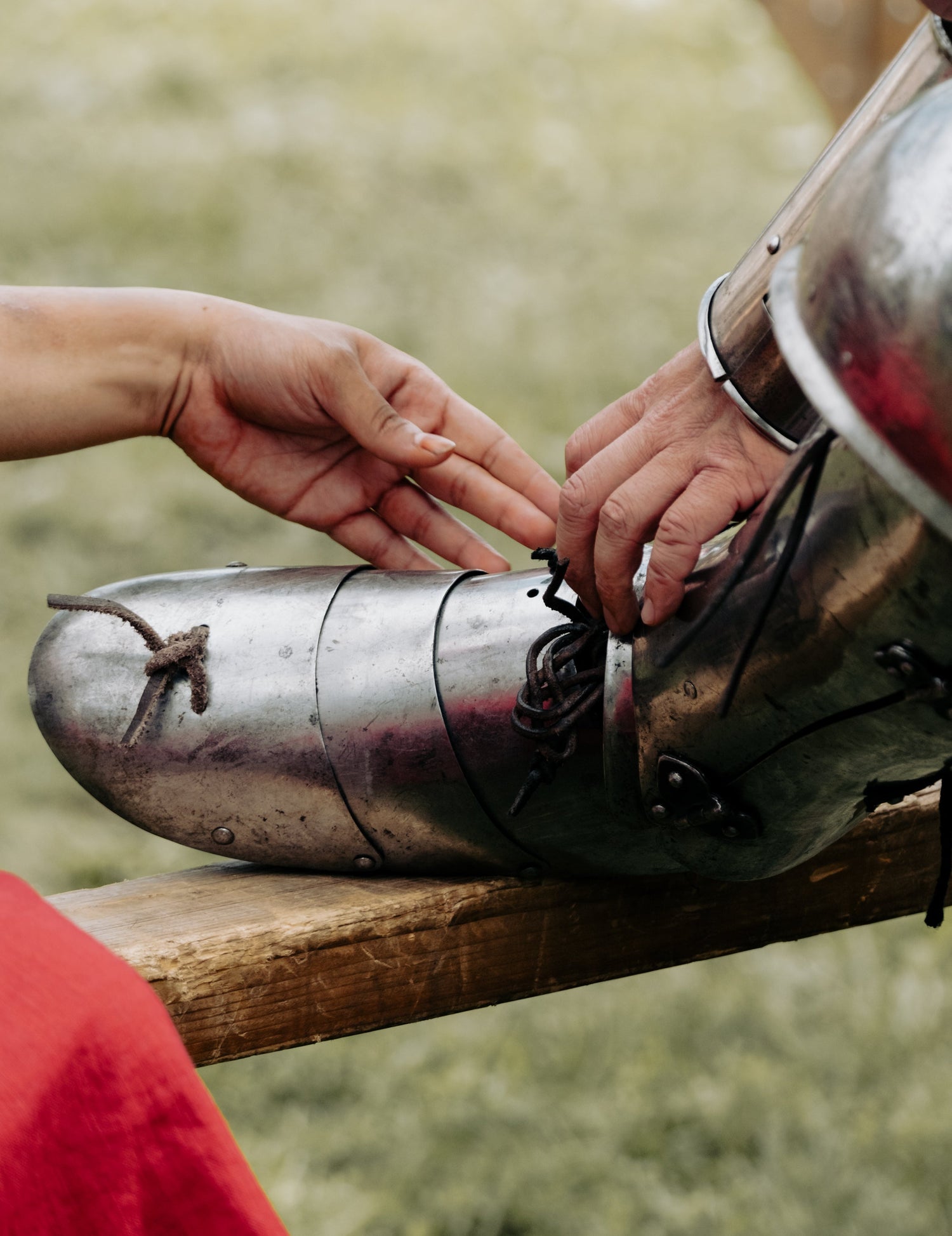 A close-up of a person's hands adjusting the laces on a polished steel medieval-style leg armor piece resting on a wooden surface outdoors. event photography finland