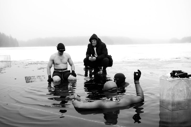 Black and white photograph of three people in a frozen body of water, with one standing shirtless in swim trunks, one crouching in a dark jacket, and one floating on their back wearing a swim cap. A large ice block is visible on the right.