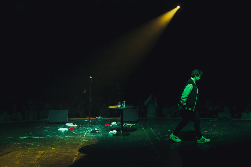 ismo leikola finland comedian. A person walks across a dark stage under a spotlight, with green and yellow stage lighting, microphones, and scattered items on the floor.