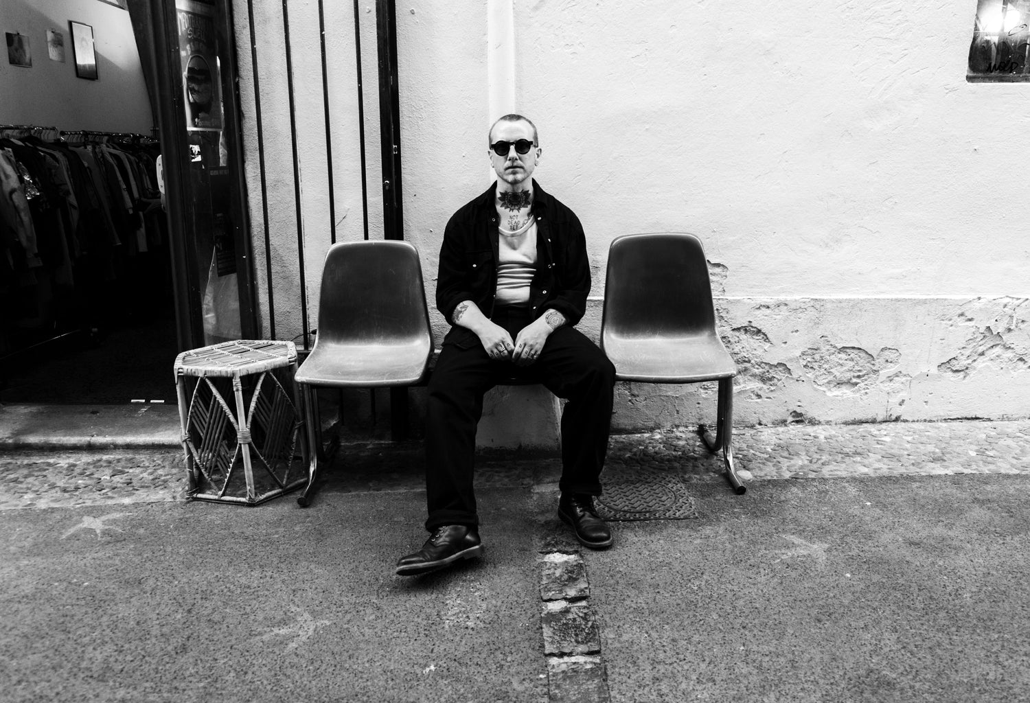 A black and white photograph of a man sitting on a bench in front of a building wall, wearing sunglasses, a dark jacket, and a striped shirt.