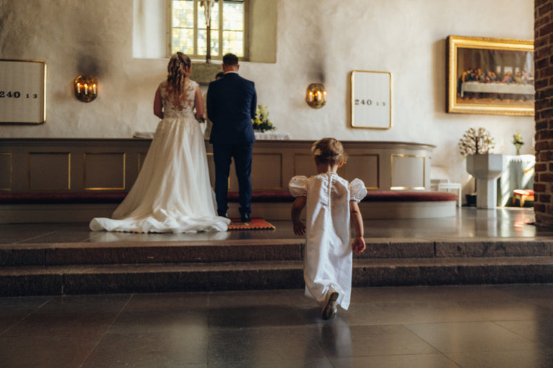 A bride in a white wedding gown and a man in a navy suit stand at an altar inside a church, while a young girl in a white dress walks toward them from the foreground on a polished floor.