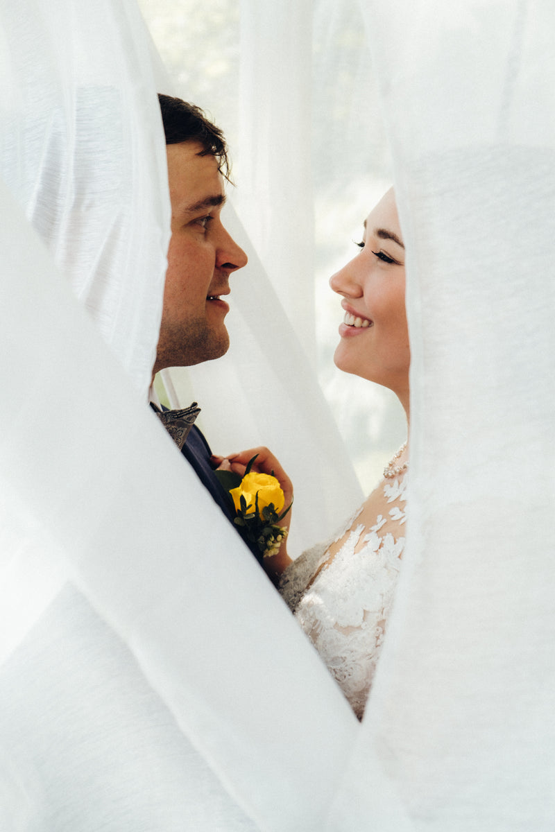 A close-up wedding portrait of a bride and groom peeking through white bridal veils. The bride is smiling and wearing a white lace dress, while the groom wears a dark suit and holds a yellow flower. helsinki finland